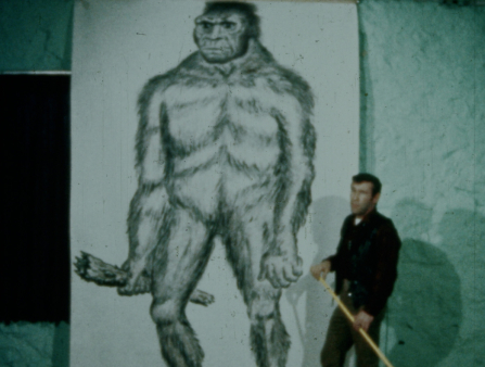 Grainy footage of a man dwarfed by a large-scale illustration of bigfoot.