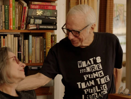 An elderly white man with thinning short white hair wears glasses and a black t-shirt that reads "What's more punk than the public library?" smiles toward an older white woman with shoulder-cropped hair and red glassed who stares at him admiringly