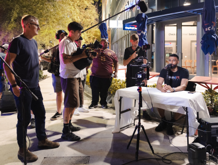 A camera crew is seen shooting a dark-skinned man with a neatly-trimmed beard who sits at a makeshift table staring into a rigged smartphone that serves as his camera