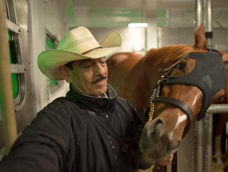 A dark-skinned man with a mustache and a cowboy hat is in a stable holding onto horse's head nearby