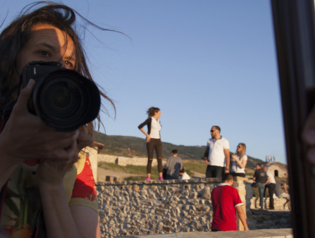 A white woman holding a camera is seen in a mirror as a group of people stand along in the background in a desert field