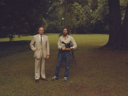 Two white men (one in a tan suit and tie; the other in casual clothes, holding a camera) stand in a field with trees behind them