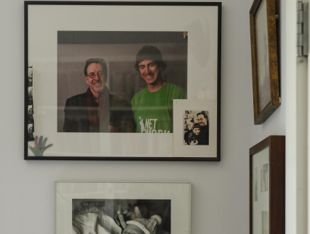 Shot of a wall of family photos, including a framed one of a middle-aged father smiling with his son above a black and white baby pic