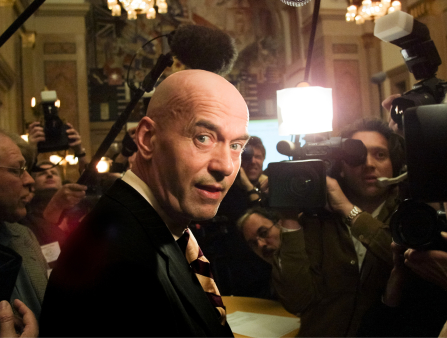 A white bald man in a suit is surrounded by media photographers and videographers as he stares back to look at the camera