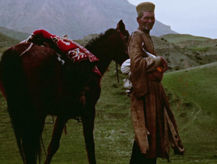 A dark-skinned older farmer stands in front of lush green mountains, a horse by his side