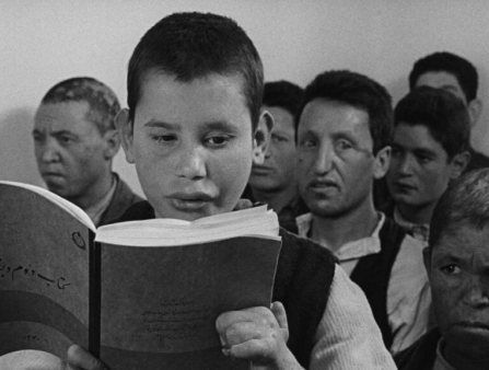 Black and white image of a young boy reading from a notebook, many others right behind him