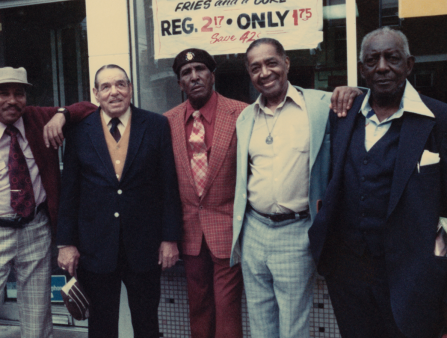 Five Black middle-aged men in sharp suits of all colors, many with colorful ties to match, stand smiling at the camera while on the street near a storefront