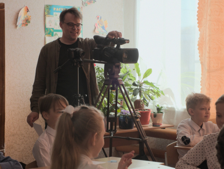 A young white man with glasses holds a camera in a classroom, surrounded by kids at their desks
