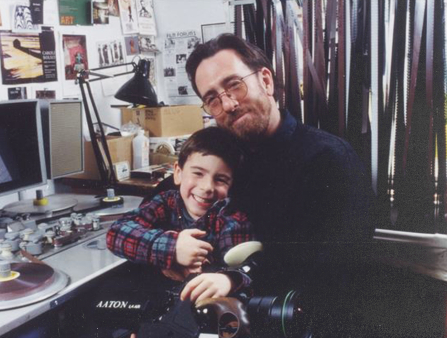 Happy photo of a middle-aged man with brown hair and beard, hugging his young son, who sits on his lap at his cluttered office