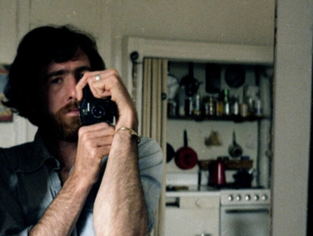 A middle-aged man with long brown hair and bears holds a camera with both his hands, capturing his reflection in a mirror, a kitchen behind him