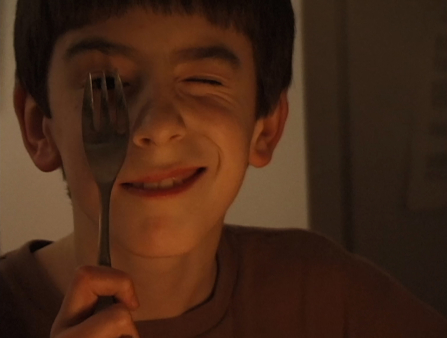 A young boy holds up a spork against his face, squinting and smiling as he does.