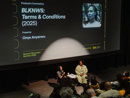Two Black women are seated in front of a crowded theater as a screen behind them reads the title of a film: BLKNWS: Terms & Conditions