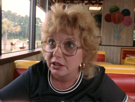 Medium shot of a heavy-set white woman wearing a ringer blue shirt, large-framed glasses and curly blond hair sitting at an old-school-looking diner