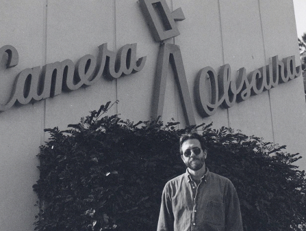 Black and white photo of a middle-aged man with dark hair and bushy beard standing in front of a building adorned with "Camera Obscura" signage