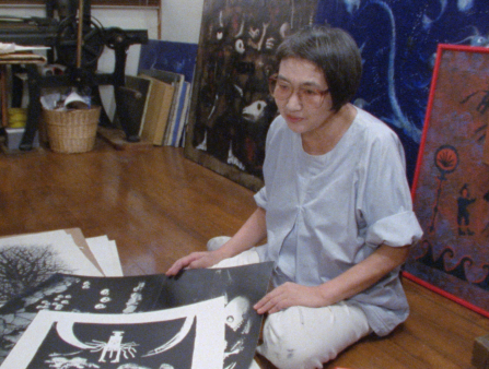 An older Asian woman with short black hair and glasses sits on the floor surrounded by paintings and artwork