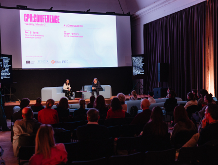A young Asian woman and a white man sit on giant sofas on a stage in front of an audience with a screen behind them that reads "CPH:CONFERENCE"