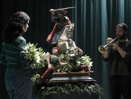 A young woman with long wavy brown hair in a striped sweater and jeans stares at a colorful sculpture of a man on a horse as a trumpet player plays in the background
