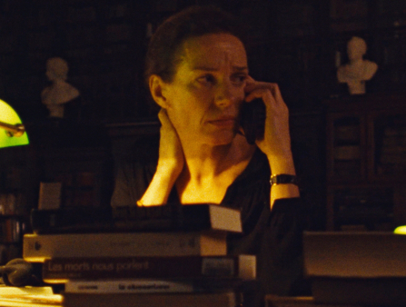 A middle aged white woman with hair pulled back holds her phone against her ear while seated at a desk, lit by a desk lamp