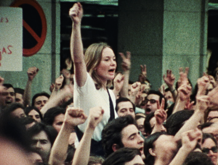 Grainy image of an outdoor protest with many men and women holding their fists up, a white blond woman rising above the crowd in a similar pose
