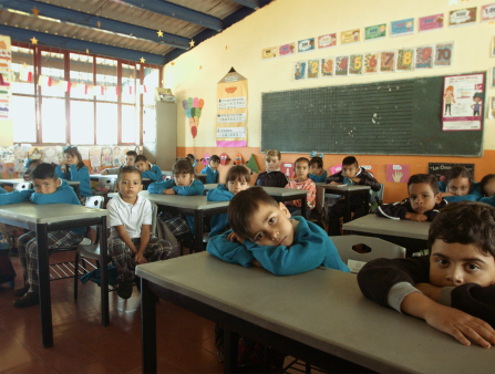 A group of dark-skinned uniformed schoolkids lay down their faces on their desks with a blackboard and kids art behind them
