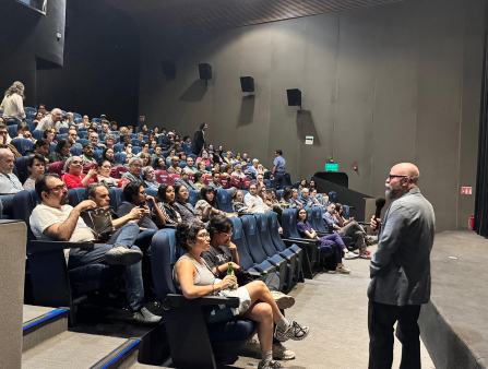 A bald white man in a grey suit holds a mic and addresses a packed theater screening