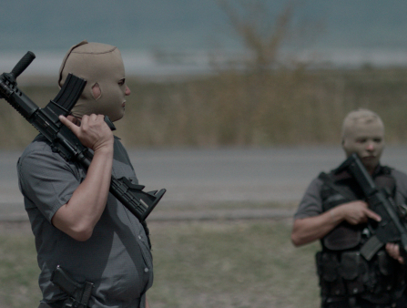 Two men in cammo gear stand in an empty field holding guns don skin-colored masks that reveal only their eyes, nose, and mouth