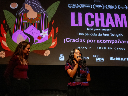 A dark-skinned woman in a colorful top holds a microphone in front of a theater screen that advertises the film Li Cham as another young woman with long brown hair in a burgundy sweater and pants stands to the side.