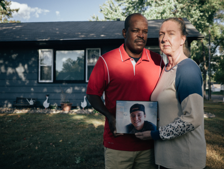 A white woman with her hair pulled back in a long-sleeved shirt poses alongside a Black man in a red polo shirt in front of a house pose holding a photo of a young boy