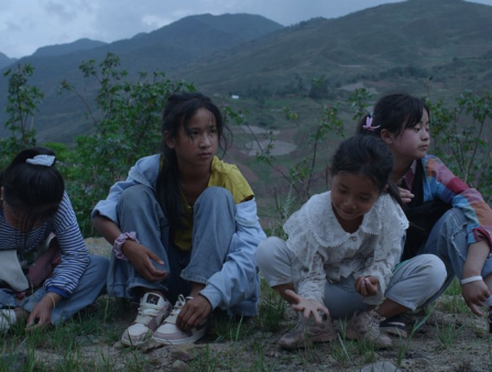 Four young Asian girls in casual clothing sit on the ground, mountains behind them