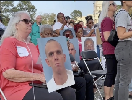 At an outdoor memorial, a woman in a red shirt and many others hold a large photo of a white man in a white tshirt.