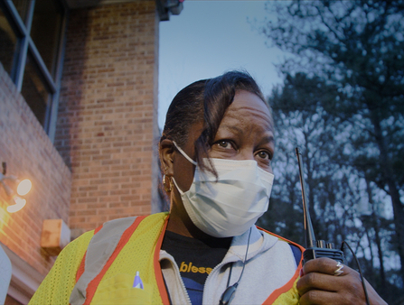A Black woman wearing a face mask and a yellow high visibility vest holds a walkie talkie.