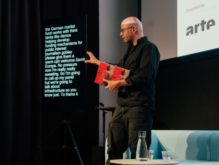 A bald man with glasses holds a CPH:DOX flyer and gestures with his right hand as he presents. A large screen appears behind him