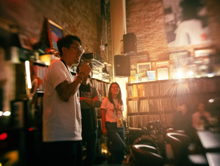 A man in glasses, a TIDF pass, and a white T-shirt speaks into a microphone. Two people stand to his left. They are in a large room with brick walls, a drum kit, large speakers, and tall shelves of records