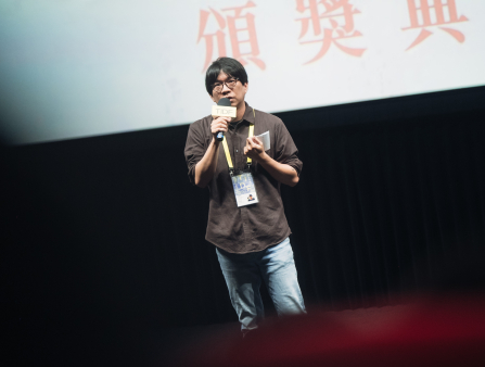 An Asian man in glasses, a brown, button down collared shirt, jeans, and a TIDF pass around his neck speaks into a microphone from a stage. The microphone is labelled TIDF. The bottom portion of a large screen can be seen behind the man, displaying Chinese characters