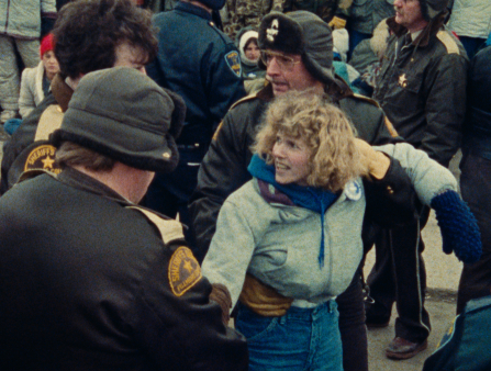 A blonde woman in a blue jacket is restrained by three men with hats and sheriff's badges. Two more officers stand behind them in addition to a crowd