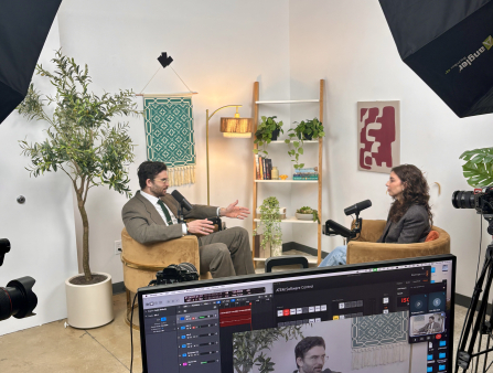 A man in a suit and glasses sits across from a woman in a grey jacket and jeans. Both are in armchairs and speak into microphones. In the foreground, a monitor can be seen displaying the man's face