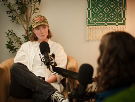 A woman in a white shirt and camo hat sits in an armchair with a microphone in front of her. The back of the head of another woman can be seen in the bottom right corner