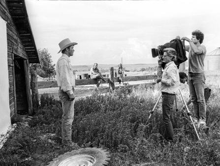 Black and white image of two men taking a photo of a man in a cowboy hat on a farm. Two onlookers sit on a wooden fence in the background