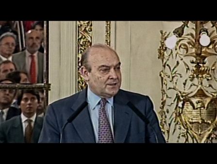 Aging balding man in suit and tie addresses an unseen crowd at a mic stand with golden decor behind him