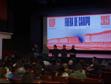 Group of men in casual clothing sit on a stage with a big movie screen behind them in front of a packed movie theater audience