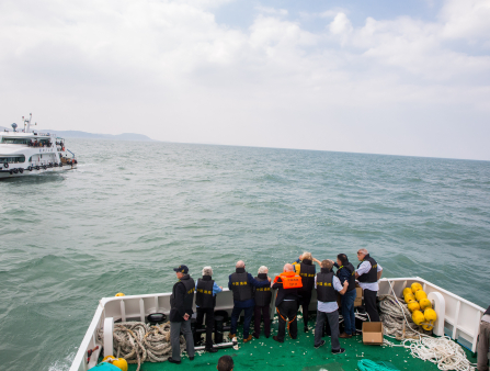 Nine figures in black life vests stand on the edge of a ship looking over the ocean. Another ship is in the background