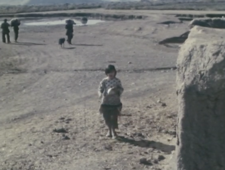 Young girl stands alone in desert looking at the camera, three people and a dog walk in the distant background