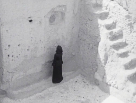 Black and white shot of figure in dark robe standing in ruins facing wall