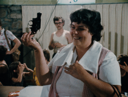 A smiling white woman, with short dark hair, a white shirt with a large collar, and a red vest, holds up a small revolver. She is inside and other people appear to be conversing behind her