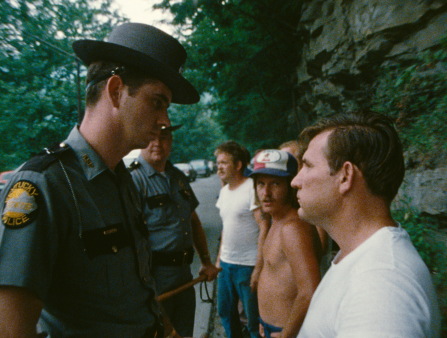 A Kentucky police officer and a shorter white man in a white T-shirt stare intensely at each other. Behind the two, another officer and three civilians, one also in a white T-shirt and one with no shirt and a baseball cap, look on