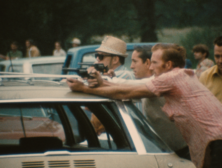 Two white men, one in a striped red button-down with the sleeves rolled up, rest revolvers on top of a car, pointing the guns and staring intently at someone or something off camera to the left. More men in the background stand behind cars, facing the same direction