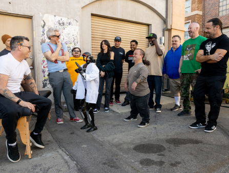 12 casually dressed adults gather around a humanoid robot wearing a lab coat in an alley