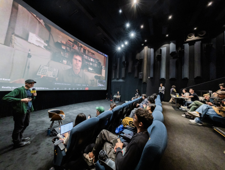 The front three rows of a theater, filled with people holding notetaking and camera equipment, look at a man in a green shirt holding a microphone and a man on the theatre screen who is labelled on Google Meet as Ersnt Karel. Karel has curly hair, glasses, and a green shirt buttoned all the way up. He sits in front of sound equipment and a shelf of CDs