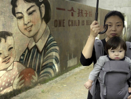 A woman holds an umbrella over herself and her young son, who sits in a harness she is wearing in front of her.  Behind the two, a large mural portrays a woman and her child and reads in both Chines and English "ONE CHILD IS BEST"