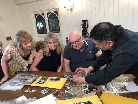 An Asian man shows a white man and two white women and image on a phone screen. Laid out on the table in front of the group are a variety of photographs and papers. The group sits in front of a wooden table with white wood panelling and a small window behind them